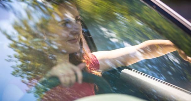 Motion blured image of happy young woman driving a car on a summer road trip travel vacations. Shot trough windshield. - Foto, immagini