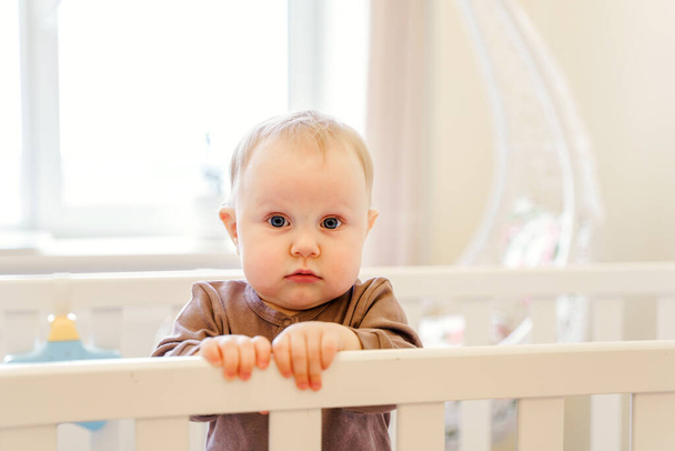 Een schattige baby houdt een wieg vast in een heldere thuiskamer - Foto, afbeelding