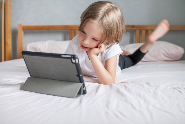 Little girl doing homework online, lying on the bed at home. Communication with relatives online. The concept of the beginning of the new school year - Фото, изображение