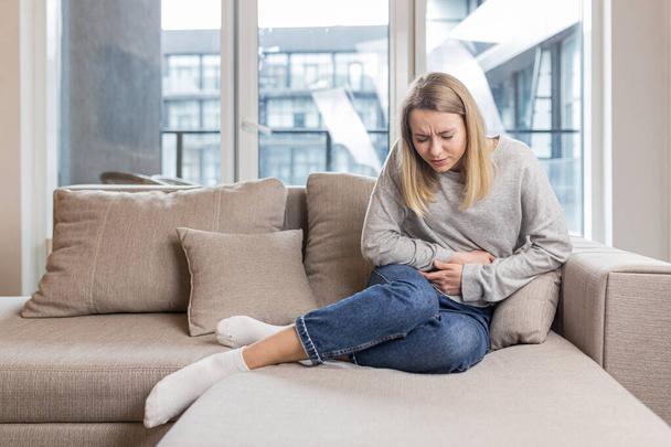 Young woman sitting on sofa holding hands on stomach twisted from pain, poisoned or menstrual pain, woman at home - Foto, Bild