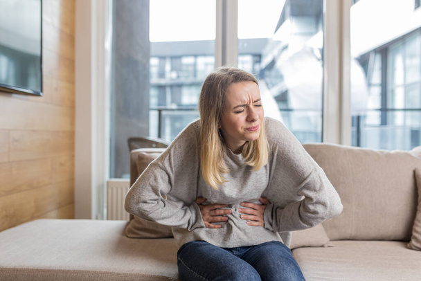 Young woman sitting on sofa holding hands on stomach twisted from pain, poisoned or menstrual pain, woman at home - Foto, Bild
