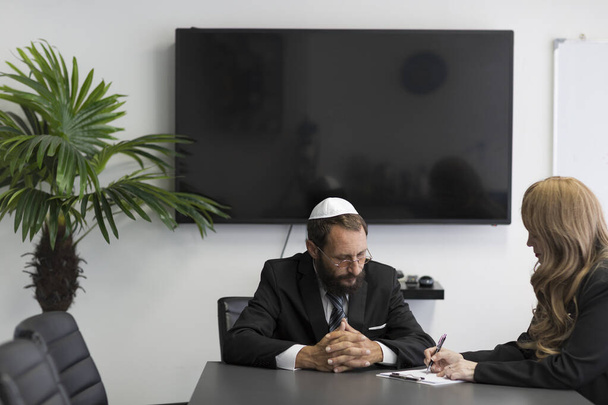事務所に座っているユダヤ人男性と眼鏡をかけ、エレガントな女性が彼女のアイデアを書き留めています。ユダヤ人男性のシャツ、スーツ、国民の帽子キプパフは事務所で一緒に働いています。男に集中しろ - 写真・画像