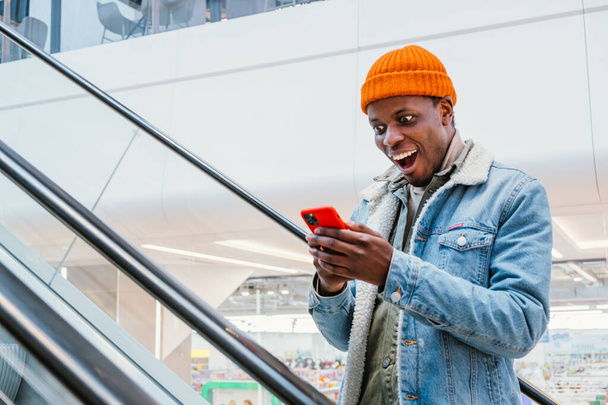 Szczęśliwy Afroamerykanin patrzy na telefon idzie w górę schody ruchome - Zdjęcie, obraz