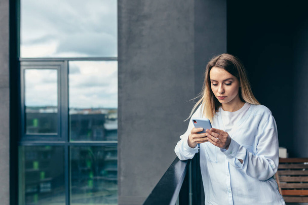 Beautiful blonde woman uses a mobile phone while standing on the balcony of an apartment building - Foto, afbeelding
