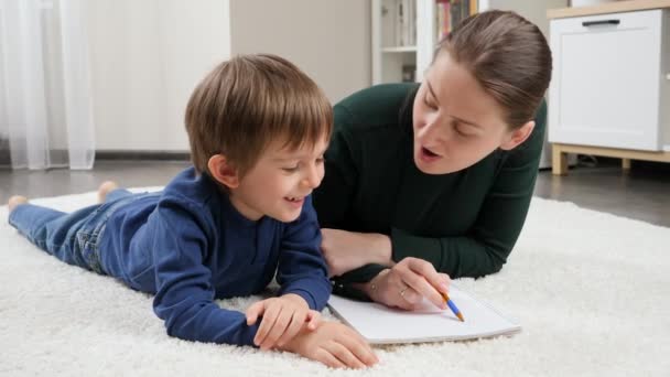 Porträt einer lächelnden Mutter, die ihrem kleinen Sohn, der im Wohnzimmer auf dem Boden liegt, das Schreiben beibringt und bei den Hausaufgaben hilft. Konzept der Kindererziehung, Entwicklung und glücklichen Elternschaft. - Filmmaterial, Video