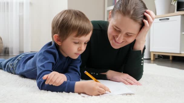 Lächelnder Junge, der auf dem Boden liegt und mit seiner Mutter Hausaufgaben macht. Konzept der Kindererziehung, Entwicklung und glücklichen Elternschaft. - Filmmaterial, Video