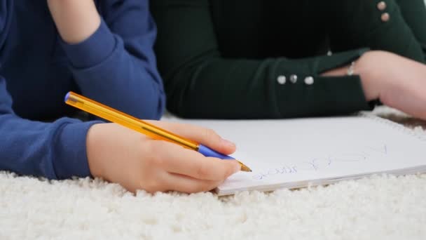 Großaufnahme eines kleinen Jungen, der mit Bleistift im Schreibheft schreibt, während er im Wohnzimmer auf dem Teppich liegt. Konzept der Kindererziehung, Entwicklung. - Filmmaterial, Video