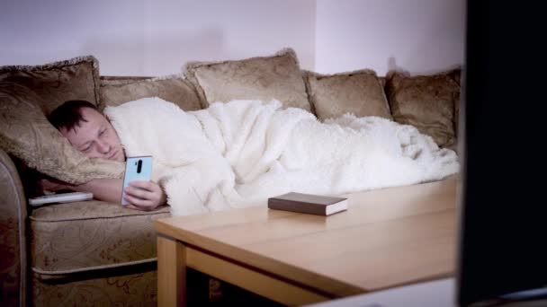 Un hombre se acuesta en el sofá por la noche viendo la televisión, sostiene un teléfono en sus manos, la iluminación de la noche - Metraje, vídeo