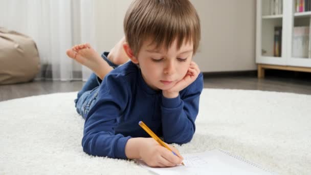 Pieni poika tekee läksyjä ja kirjoittaa copybookissa lattialla kotona. Kotimaisen koulutuksen ja lasten kehityksen käsite. - Materiaali, video