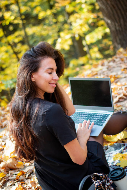 A brunette woman is sitting in an autumn park with a laptop and a cup of coffee. Student. Teacher. Online education. - Zdjęcie, obraz