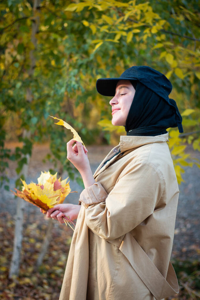 A Muslim woman in a hijab in a beige trench coat stands with autumn leaves. Close up. Black cap. - Photo, Image