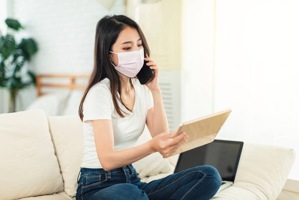 Young woman working at home, wearing a mask to protect herself, sitting on the sofa using a laptop to telecommute, and talking on the phone while watching the tablet - Photo, Image