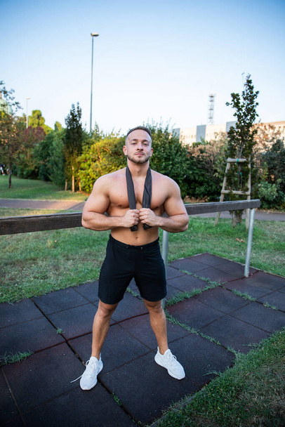 Full length of a male bodybuilder in an outdoor gym working out, shirtless, has a satisfied expression and it's summer. - Φωτογραφία, εικόνα