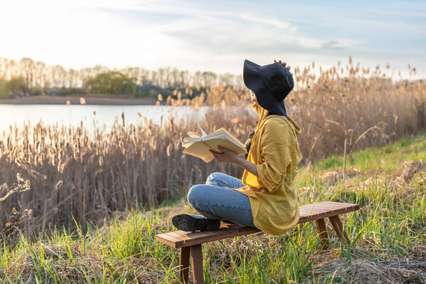 Привлекательная девушка в шляпе читает книгу на природе на закате. - Фото, изображение