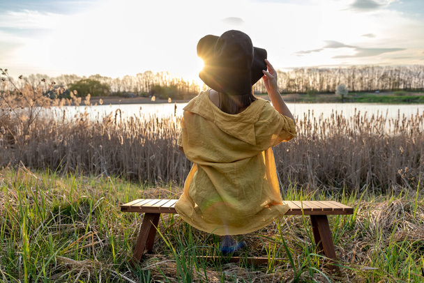 A girl in a hat and in casual style sits on a bench near the lake at sunset. - Photo, Image