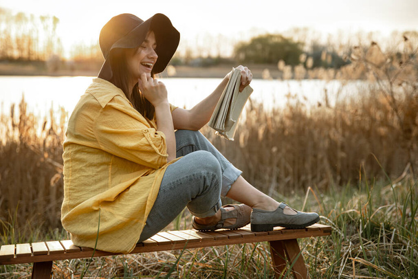 Młoda kobieta w kapeluszu z uśmiechem na twarzy czyta książkę siedzącą nad rzeką o zachodzie słońca.. - Zdjęcie, obraz