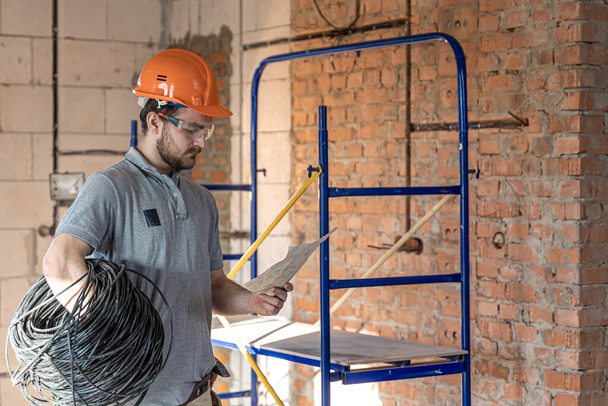 Bir elektrikçi, şantiyede elinde bir elektrik kablosu tutarken bir inşaat çizimini inceliyor.. - Fotoğraf, Görsel