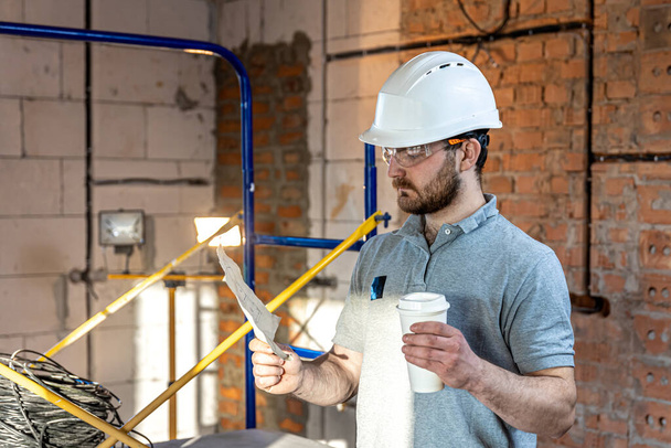 Un electricista está estudiando un dibujo de construcción con un café en la mano. - Foto, Imagen