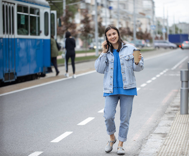 Street portrait of a cheerful young woman talking on the phone with coffee to her hand. - Photo, Image
