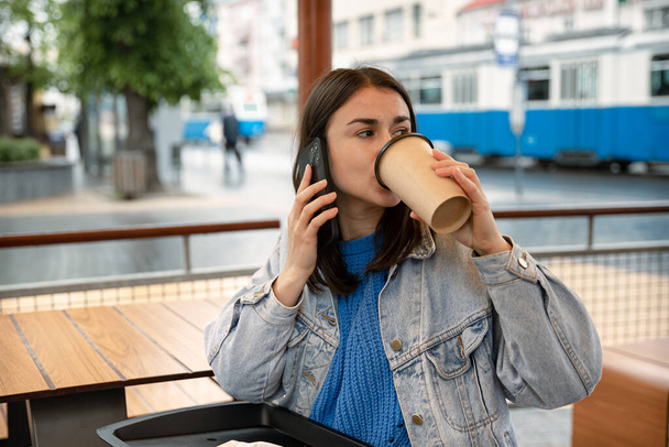 Egy fiatal nő utcai portréja, aki kávét iszik, telefonon beszél és vár valakire.. - Fotó, kép