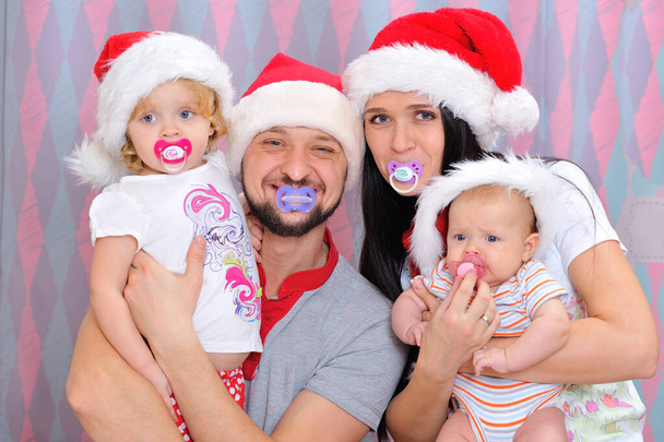 クリスマスだ。父親、母親、赤ちゃんの女の子と新生児の家族の肖像画。4人とも口に乳首と赤いクリスマスの帽子を頭にかぶってる - 写真・画像
