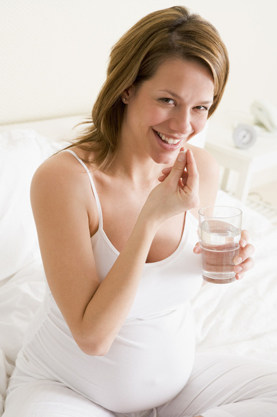 Pregnant woman in bedroom taking medicine holding water smiling - Photo, Image