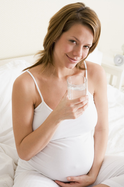 Pregnant woman sitting in bedroom with glass of water smiling - Photo, Image