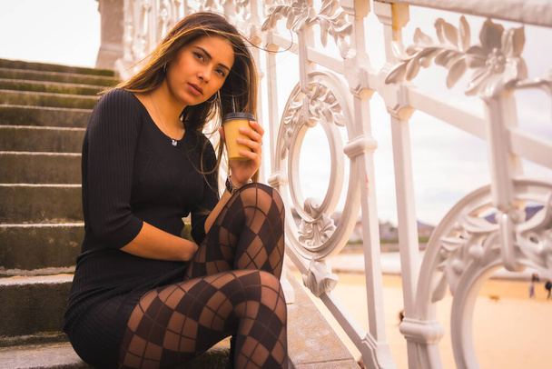 Lifestyle in the city, Caucasian brunette in a short black dress and with plaid tights sitting enjoying the vacation in summer - 写真・画像