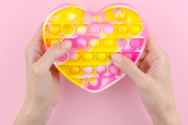 Pop es in Form eines Herzens auf einem rosa Hintergrund. Modernes Kinderspielzeug lässt es in weiblichen Händen zappeln. Silikonspielzeug zur Entwicklung der Feinmotorik. - Foto, Bild