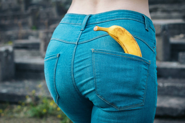 Beautiful slim ass girl in blue jeans from behind. Yellow banana in pocket booty close up. Sexy figure woman back view. Concept of sexual health, erotic, sex education - Foto, afbeelding