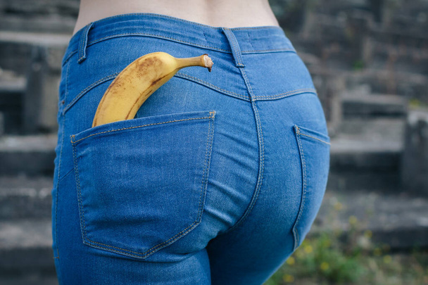 Beautiful slim ass girl in blue jeans from behind. Yellow banana in pocket booty close up. Sexy figure woman back view. Concept of sexual health, erotic, sex education - 写真・画像