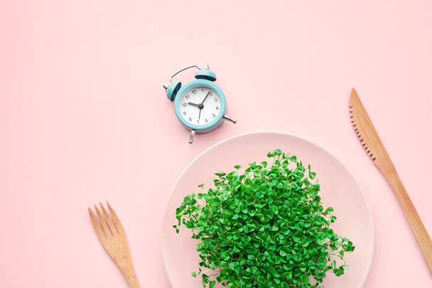 Alarm clock, cutlery and plate with greenery on pink. Intermittent fasting, lunchtime and dieting concept. - Photo, Image