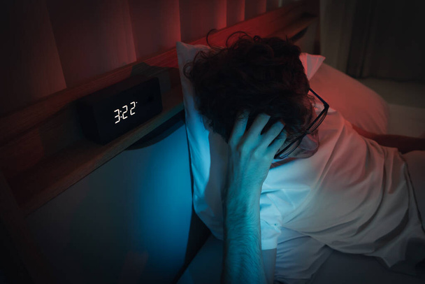 不眠症の眠れない男が問題を抱えて夜遅く目覚まし時計と彼のベッドの上で夜遅くに伝える. - 写真・画像