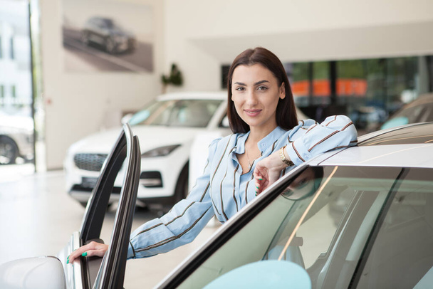 Joven hermosa mujer sonriendo a la cámara mientras elige el coche para comprar en el concesionario - Foto, Imagen