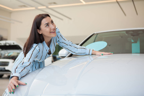 Aufgeregte junge Frau umarmt ihr neu gekauftes Auto im Autosalon, Kopierraum - Foto, Bild