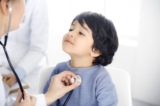 Doctor-woman examining a child patient by stethoscope. Cute arab boy at physician appointment. Medicine concept - Fotó, kép