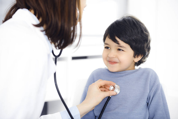 Doctor-woman examining a child patient by stethoscope. Cute arab boy at physician appointment. Medicine concept - Zdjęcie, obraz