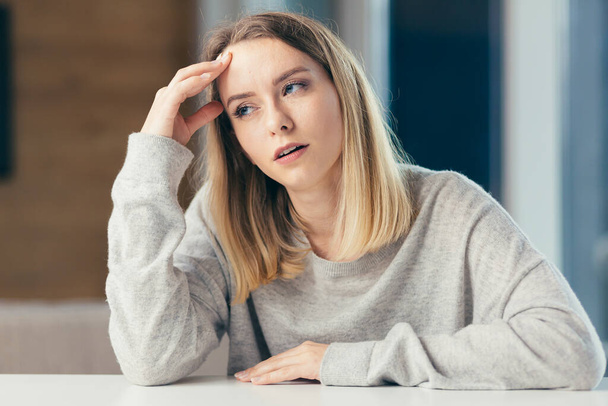 Portrait of a young blonde woman sitting at a table in the office or at home and holding his head because of severe pain. Headache or migraine or premenstrual fever indoors. In casual clothes - Photo, Image