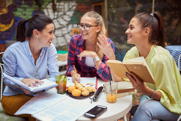 three young adult caucasian women studying together in outdoor cafe, smiling, laughing, talking - Foto, afbeelding