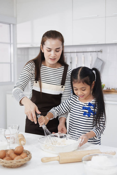 笑顔アジアの母親と小さなアジアの女の子の子供はパン屋を焼き、台所で一緒にボウルに小麦粉と卵を混ぜます。自家製のペストリーとパンのための混合成分。家族の愛と家庭学校のコンセプト. - 写真・画像