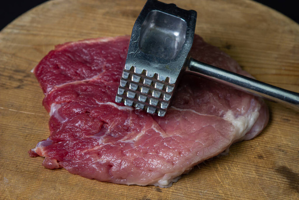 Ένα σφυρί κουζίνας και ένα κομμάτι κρέας δίπλα του. Μεταλλικό σφυρί κουζίνας για κρέας. Κρέας σε μαύρο φόντο - Φωτογραφία, εικόνα