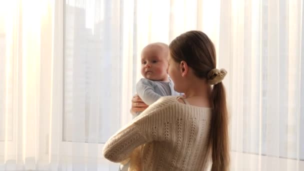 Cute little baby boy hugging his smiling mother against big window and sunset light. Concept of family happiness and parenting - Footage, Video