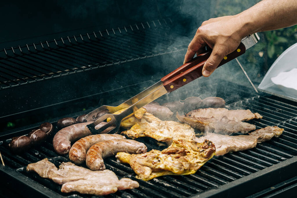 Man grillen vlees op een barbecue verschillende soorten vlees, kip, worsten, bloedworst, lam op een crbon grill in de tuin - Foto, afbeelding