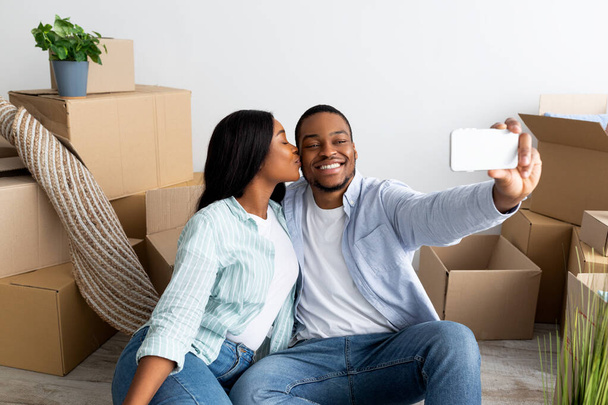 Loving black spouses moving to their own home, taking selfie among carton boxes, wife kissing husband in cheek - Valokuva, kuva
