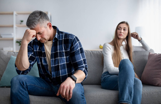 Dospělý muž v depresi sedí na gauči, hluboce zamyšlený po hádce se svou přítelkyní, uražená dáma se na něj dívá - Fotografie, Obrázek