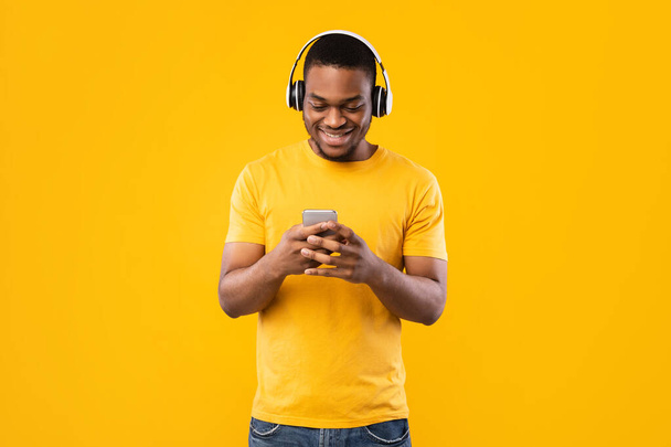 Happy Black Guy Using Smartphone Wearing Headphones Over Yellow Background - Foto, immagini
