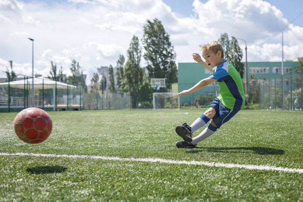 Μικρό αγόρι σε μπλε και πράσινη μορφή που παίζει ποδόσφαιρο σε ανοιχτό γήπεδο στην αυλή, ένας νεαρός ποδοσφαιριστής - Φωτογραφία, εικόνα