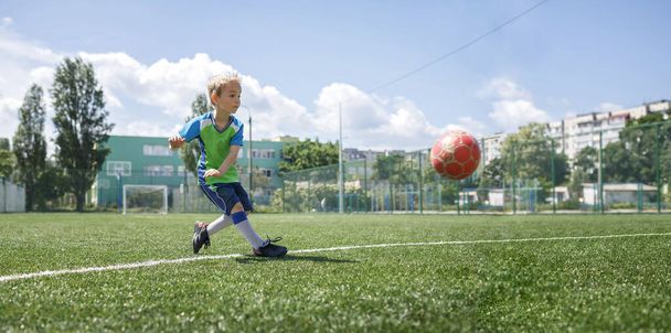 庭のオープンフィールドでサッカーをしている青と緑のフォームの小さな男の子、若いサッカー選手 - 写真・画像