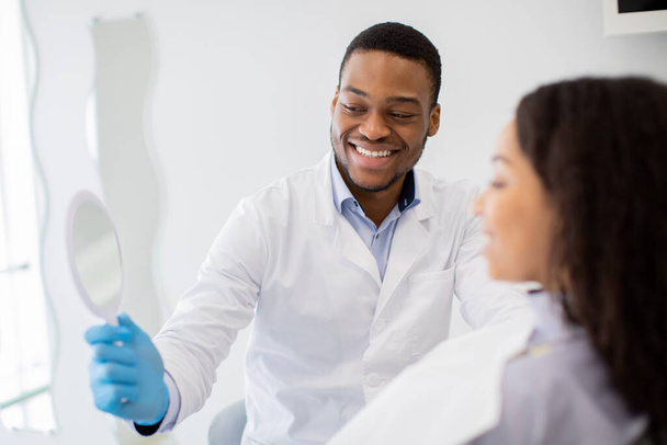 Μαύρος οδοντίατρος γιατρός κρατώντας καθρέφτη, που δείχνει το αποτέλεσμα της θεραπείας των δοντιών στον ασθενή - Φωτογραφία, εικόνα