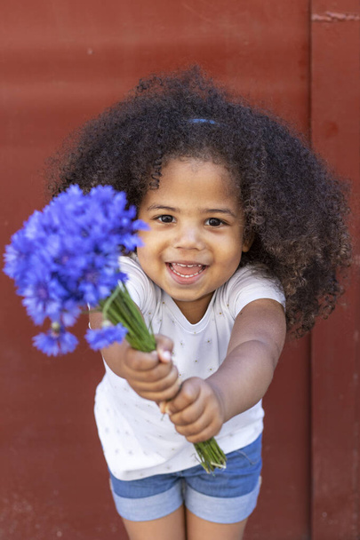 Außenporträt eines lockigen afrikanisch-amerikanischen Mädchens mit einem Strauß blauer Blumen. - Foto, Bild